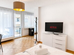 A hotel room with a bed, a TV displaying 'NETFLIX', a coat rack, and a view of the outside through large windows.