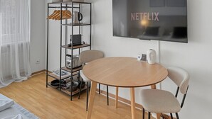 A modern room with a round wooden table, two chairs, a TV mounted on the wall, and a metal shelving unit with various items. - Lovely studio near center - Crown 1 (Zürich)