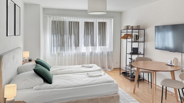 A modern hotel room with a large bed, bedside tables, a TV, a dining area, and a window with sheer curtains. - Lovely studio near center - Crown 1 (Zürich)