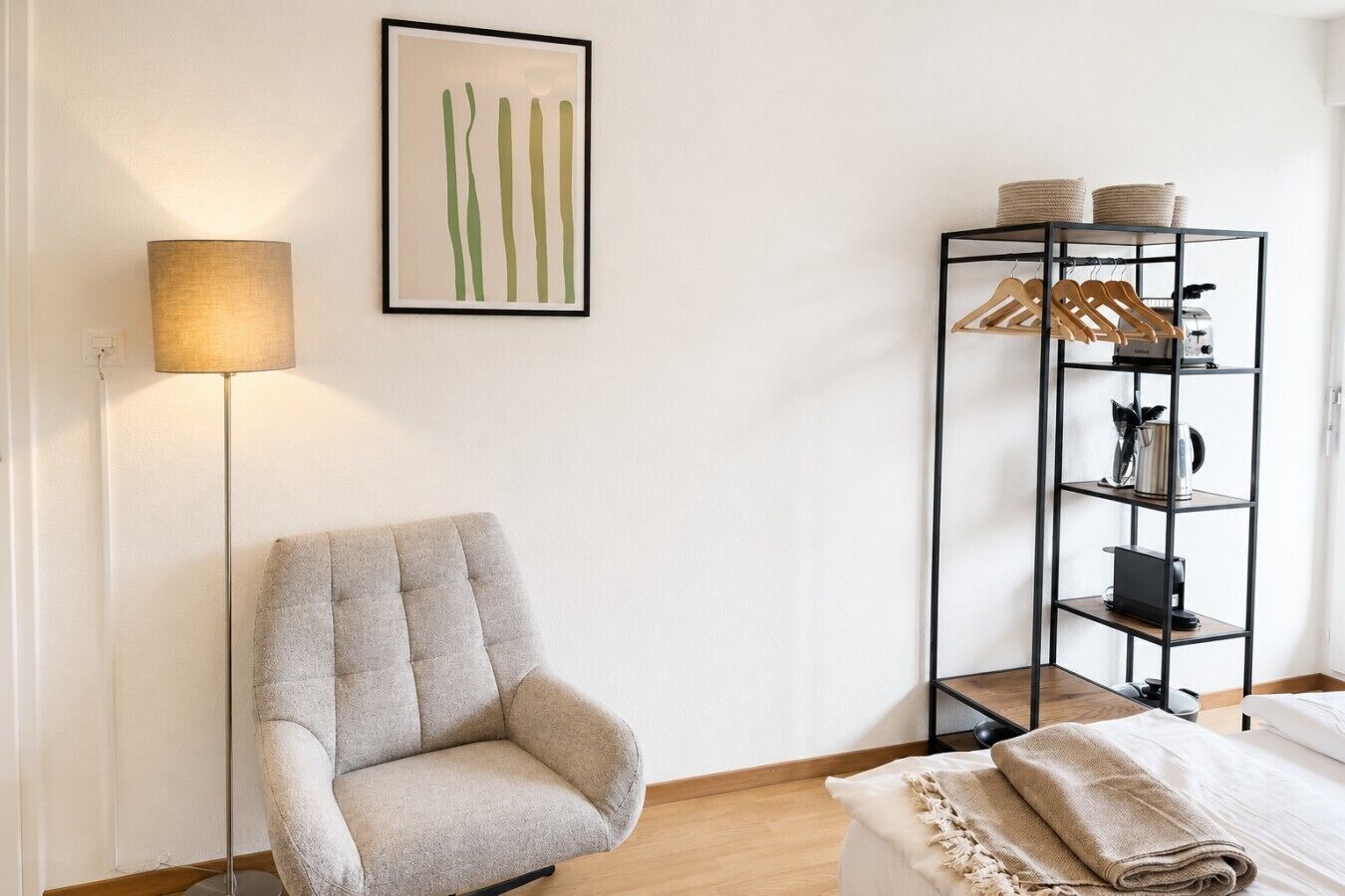A room with a chair, a floor lamp, a framed artwork, and a shelving unit with baskets and a kettle.