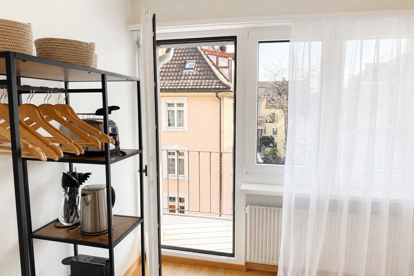 A balcony with a view of a building, a black metal shelf with hangers, a kettle, and a radiator.