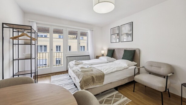 A modern bedroom with a large bed, a chair, a table, and a view of the city through the window. - City-studio on top floor - Crown 48 (Zürich)