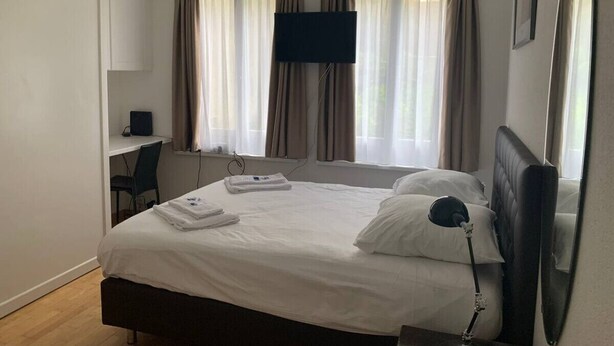 Quarto de hotel com cama, escrivaninha, cadeira, televisão e cortinas.