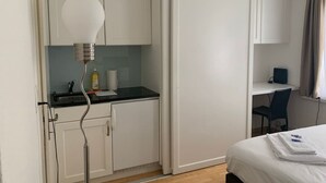 Quarto de hotel pequeno com cozinha compacta, uma cama, escrivaninha e uma cadeira.
