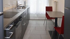 A modern kitchen with stainless steel appliances, a dining area with red chairs, and a large window with sheer curtains.