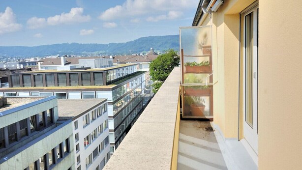 A balcony with a view of neighboring buildings and a distant mountain range.