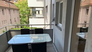 A balcony with a glass table and two chairs, a railing, and a view of neighboring buildings.