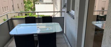 A balcony with a glass table and two chairs, a railing, and a view of neighboring buildings.