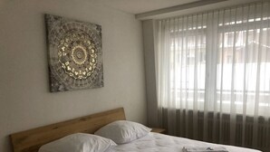 A neatly made bed with white linens and pillows, a wooden headboard, a wall-mounted artwork, and a window with sheer curtains.