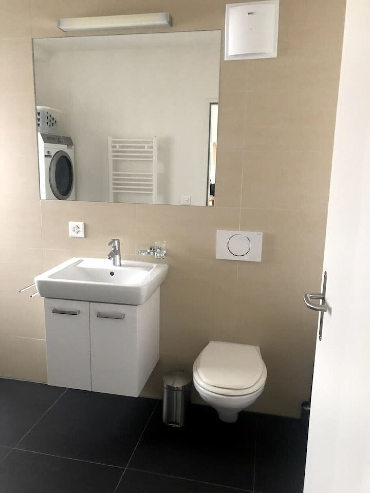 A modern bathroom with a white sink, mirror, and toilet. There is a trash can and a laundry machine in the background.