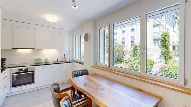 A modern kitchen with white cabinets, a built-in oven, and a dining area with a wooden table and chairs. Large windows provide a view of the surrounding buildings and greenery. - Amazing big studio city center (Sun 7) (Zürich)