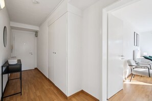 A spacious room with a wooden floor, white walls, and a large wardrobe. There is a small table with a basket on it and a chair in the corner.