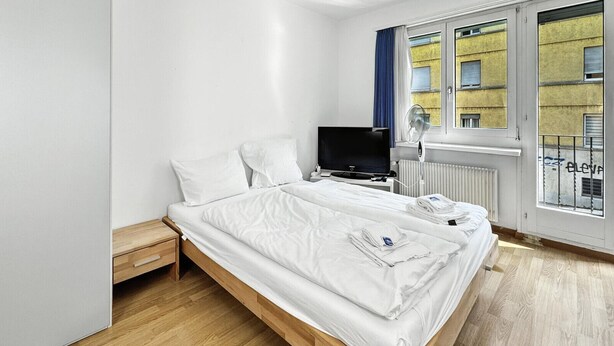 Una habitación de hotel moderna con una cama individual, televisor, mesita de noche y ventana con vistas a edificios.