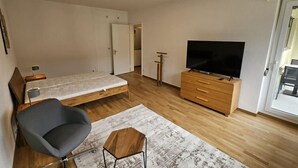 A compact room with a bed, a chair, a TV on a wooden cabinet, and a door leading to another room. - Cozy studio near train station (Wille 7) (Zürich)