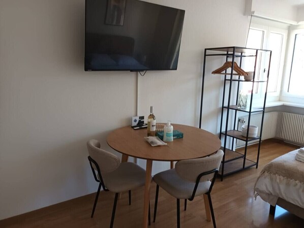 A room with a round wooden table, two chairs, a TV mounted on the wall, a radiator, and a clothing rack. - Smart studio in the city center (Crown 46) (Zürich)