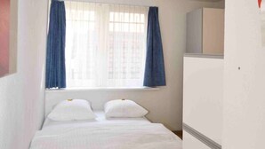 Een kleine kamer met een enkel bed, een houten vloer, witte muren en een raam met blauwe gordijnen.