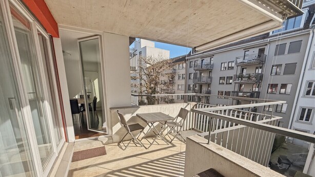 A balcony with a table and chairs, a view of neighboring buildings, and a door leading to an interior space. - 2BR near lake (Flower 23) (Zürich)