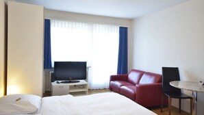Een hotelkamer met een bed, een televisie op een standaard, een rode leren bank en een kleine tafel met een lamp.