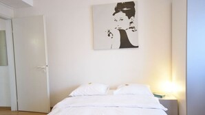 Een slaapkamer met een bed, wit beddengoed, een nachtkastje met een lamp en een wanddecoratie.