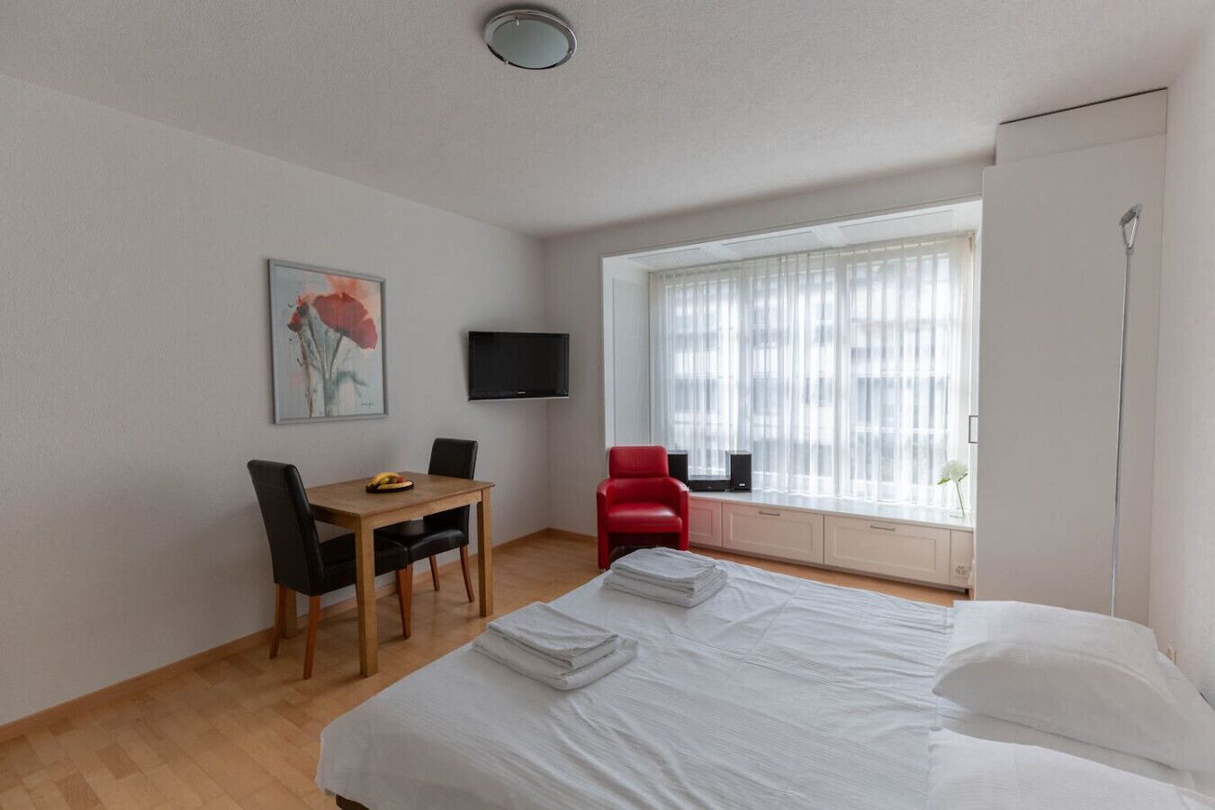 Hotellihuone, jossa on sänky, työpöytä tuolilla, punainen tuoli, televisio ja ikkuna, jossa on verhot.