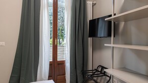 A room with a bed, a TV mounted on a white shelf, a chair, and a window with curtains.