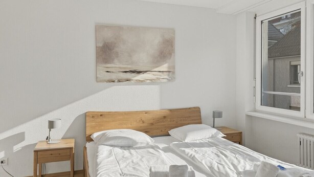 Ein Schlafzimmer mit einem Holz-Kopfteil, weißer Bettwäsche, einem Nachttisch mit Lampe, einem Fenster mit Ausblick und einem Bild an der Wand.