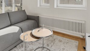 Ein modernes Wohnzimmer mit einem grauen Sofa, zwei runden Tischen, einer Heizung und Blick ins Freie durch ein Fenster.