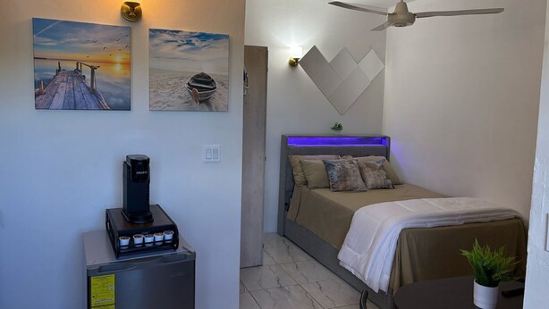 Une chambre à coucher de taille réduite, comprenant un lit, un ventilateur de plafond, une machine à café et des œuvres d’art accrochées au mur.