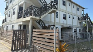 Un immeuble à plusieurs étages, doté d’un escalier menant aux étages supérieurs, d’une entrée fermée par un portail et d’une zone délimitée par une clôture où se trouve un arbuste.