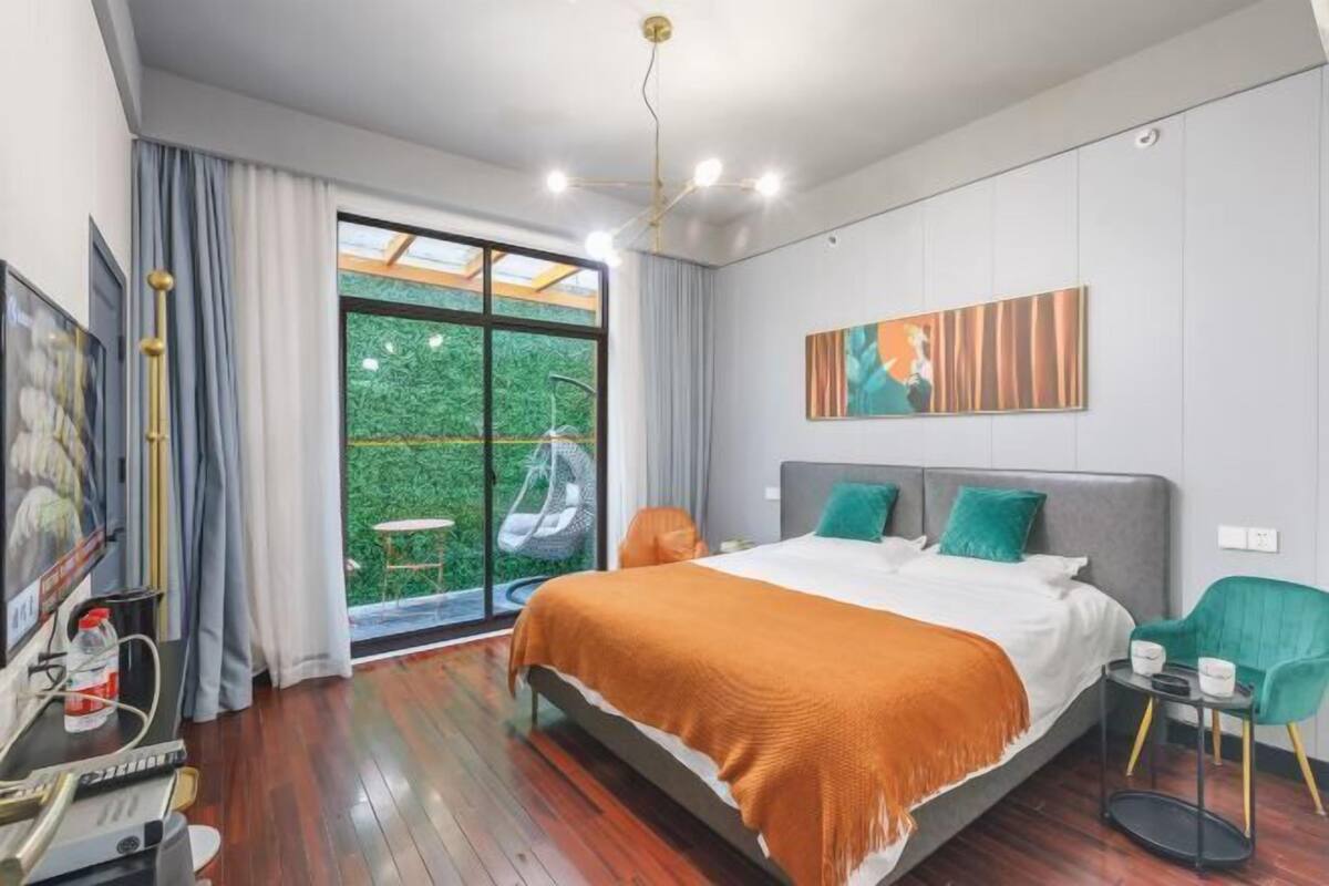 A modern hotel room with a large bed, a desk with a computer, and a view of greenery through the window.