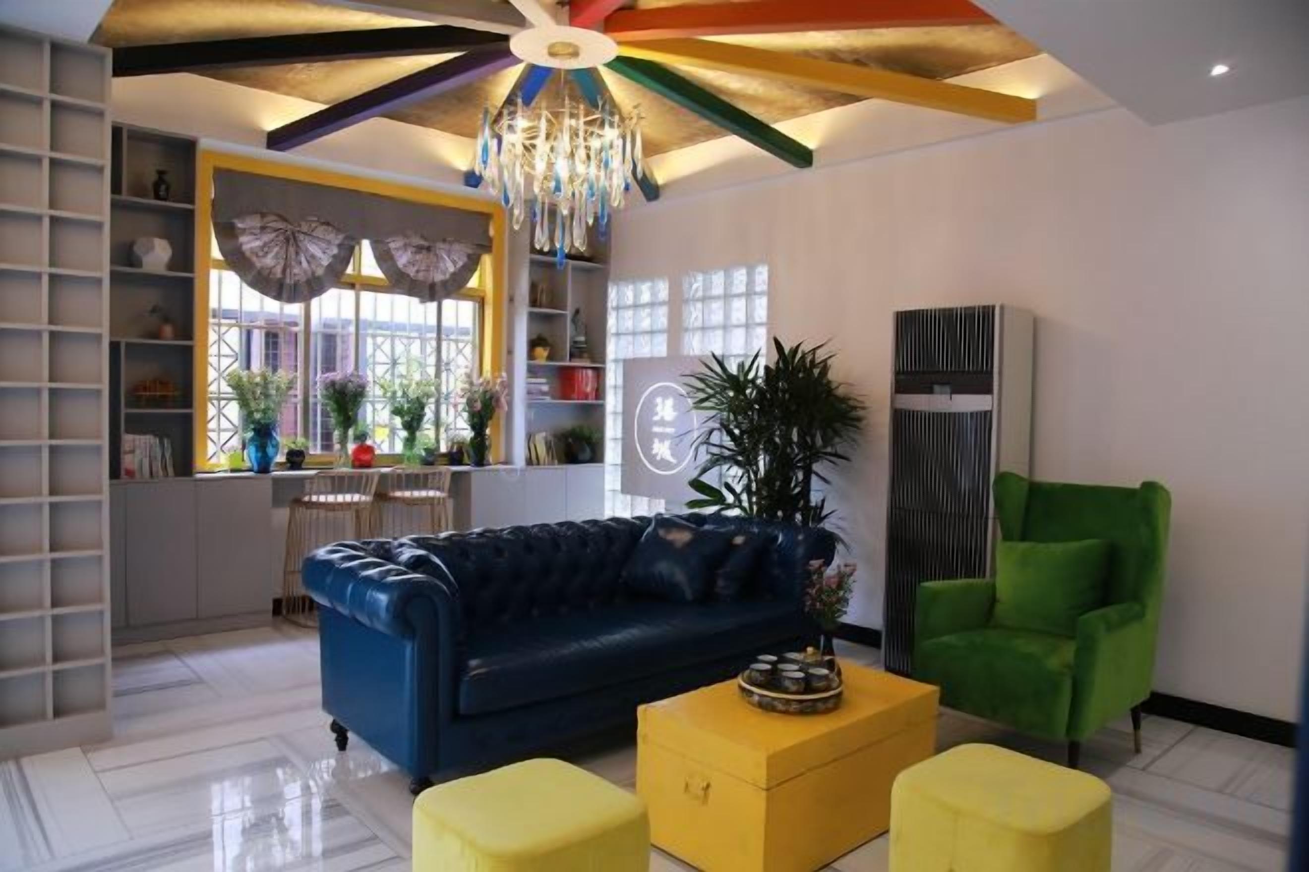 Una sala de estar con un sofá azul, un taburete amarillo y una silla verde. Hay una araña, una ventana con cortinas y una planta.