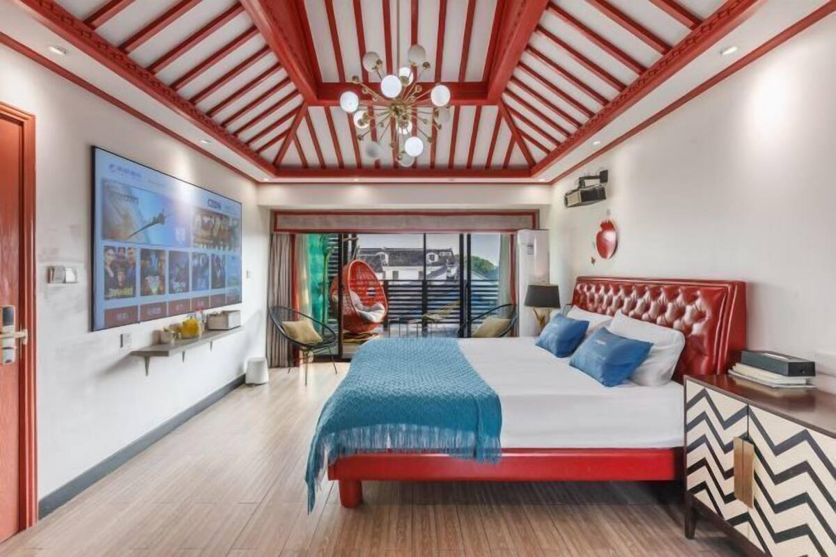 A bedroom with a bed, a red headboard, a chandelier, a wall-mounted TV, and a window with a view.