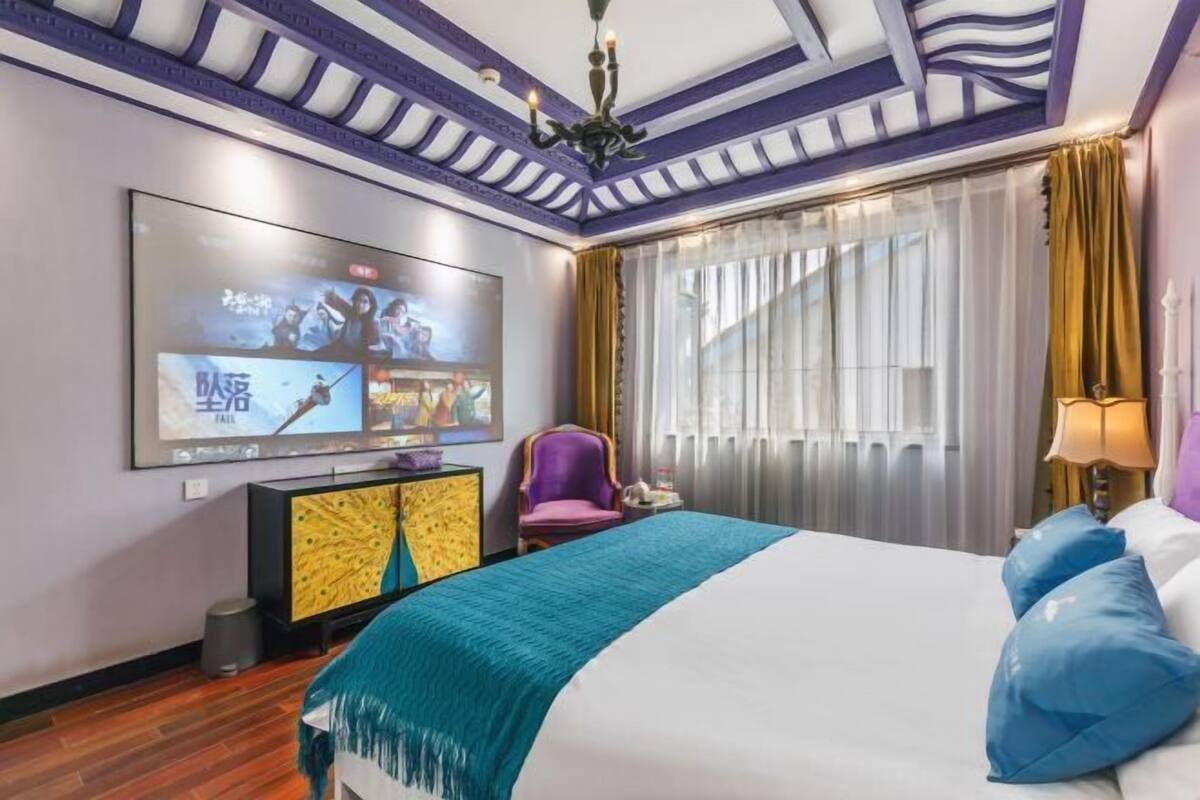 A hotel room with a large bed, a television mounted on the wall, a purple armchair, and a window with curtains.