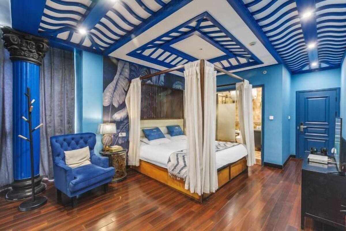 A bedroom with a four-poster bed, a blue armchair, and a ceiling with a geometric pattern.