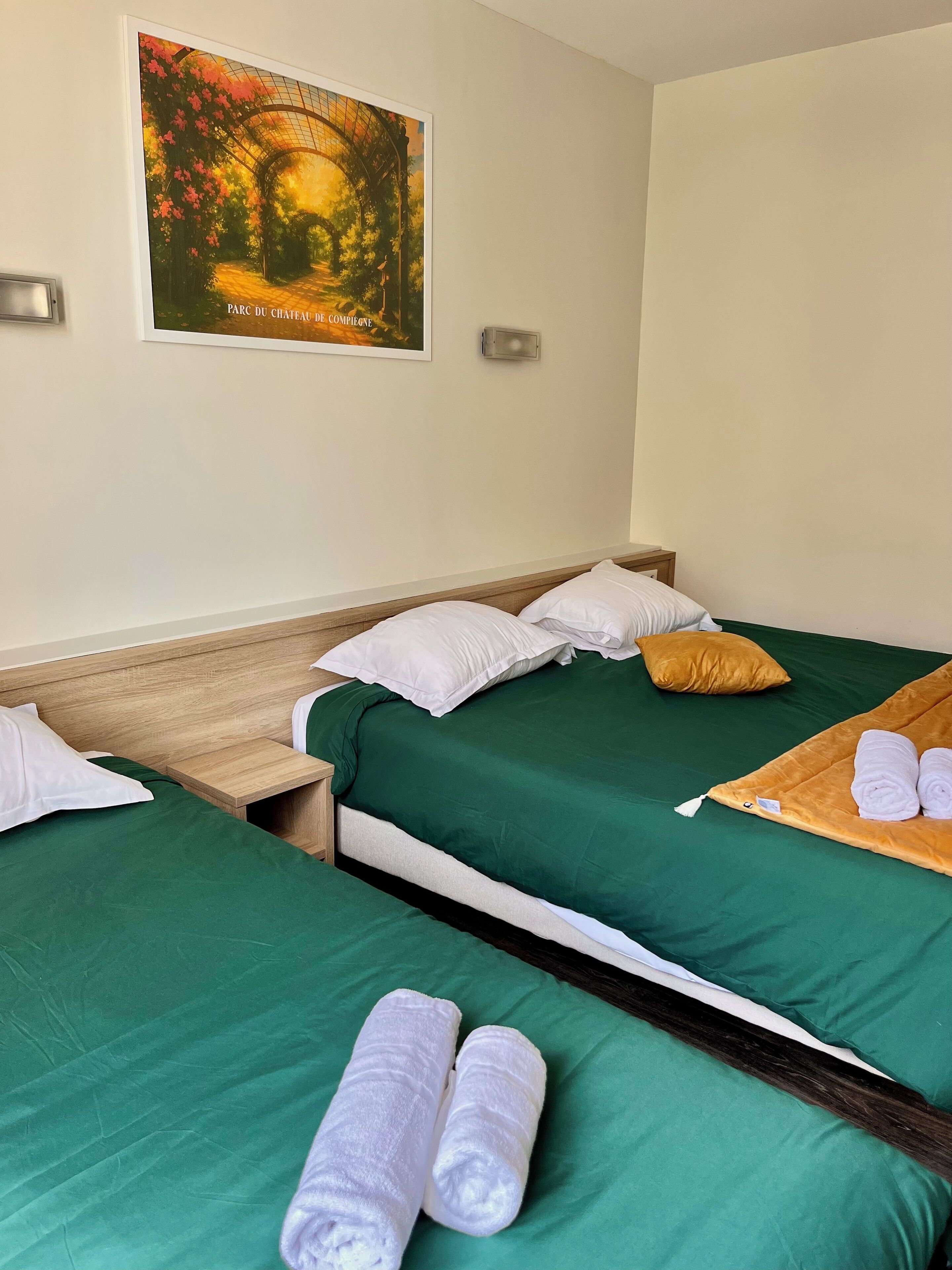 Habitación de hotel con dos camas, ropa de cama verde, almohadas blancas, cabecero de madera, mesita de noche de madera, lámpara fijada a la pared e imagen enmarcada de un jardín.