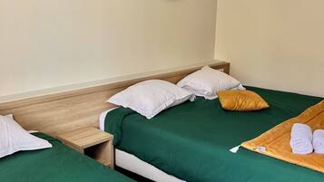 Habitación de hotel con dos camas, ropa de cama verde, almohadas blancas, cabecero de madera, mesita de noche de madera, lámpara fijada a la pared e imagen enmarcada de un jardín.
