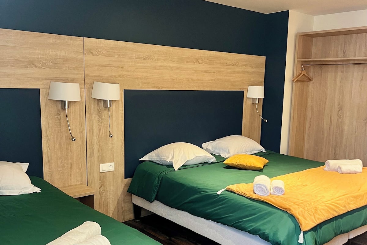 A hotel room with two beds, each with green bedding, white pillows, and a wooden headboard. There are two bedside tables with lamps, a wooden wardrobe, and a wooden bench.