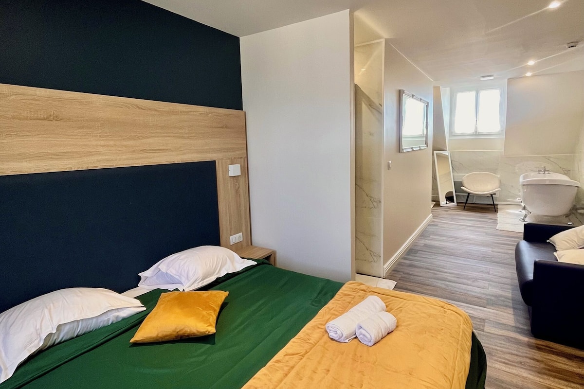 A hotel room with a bed, a green bedspread, a wooden headboard, a yellow pillow, and a bathroom with a bathtub and a mirror.