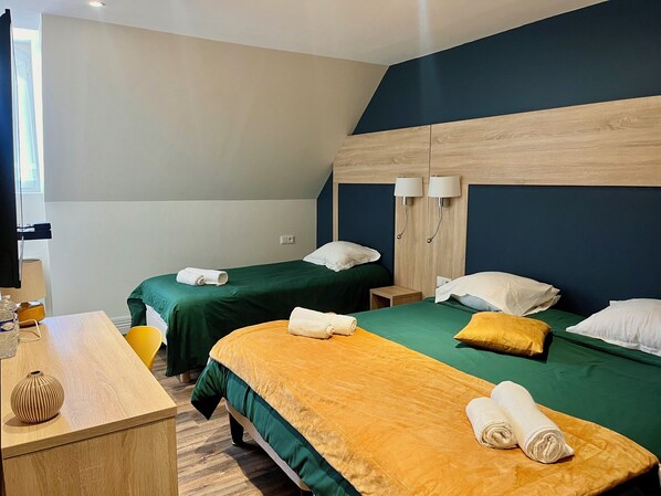 Une chambre d’hôtel avec deux lits, chacun recouvert d’une couverture verte et agrémenté d’un oreiller jaune. Il y a deux tables de chevet, l’une avec une lampe et l’autre avec une petite table de nuit en bois. On peut voir, au premier plan, un bureau en bois avec un vase et une lampe.