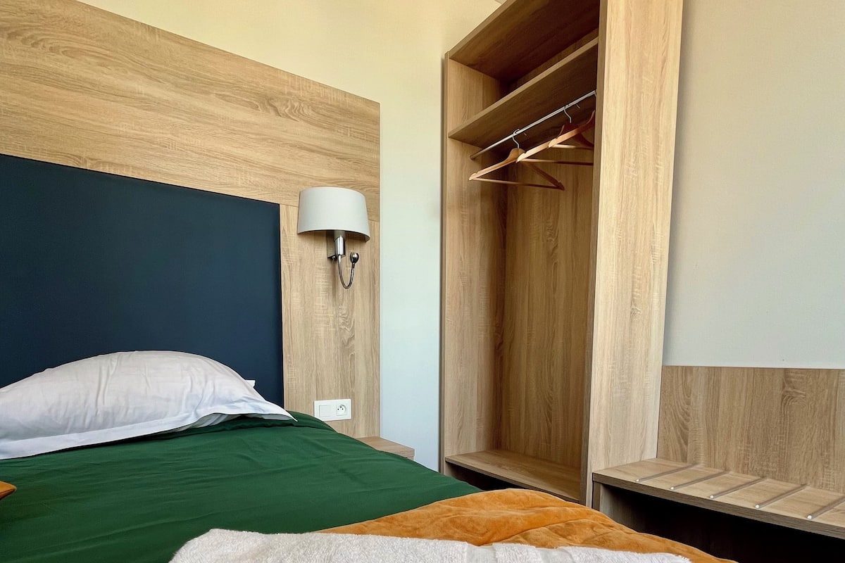 A hotel room with a bed, green and orange bedding, a wooden headboard, a bedside lamp, and a wardrobe.