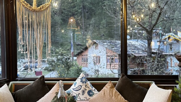 Una habitación acogedora con un ventanal grande, una lámpara colgante de macramé y una mesa con cojines y un pequeño adorno.
