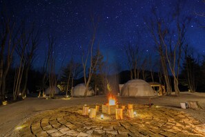 A starry night sky with a campfire in the foreground, surrounded by bare trees and covered structures.