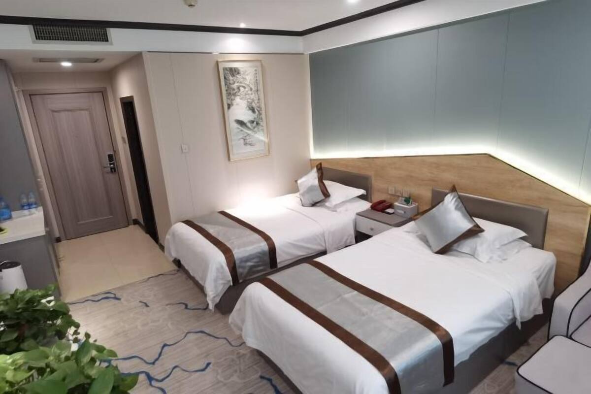 A hotel room with two beds, a bedside table with a lamp, a wall-mounted artwork, and a door leading to another room.