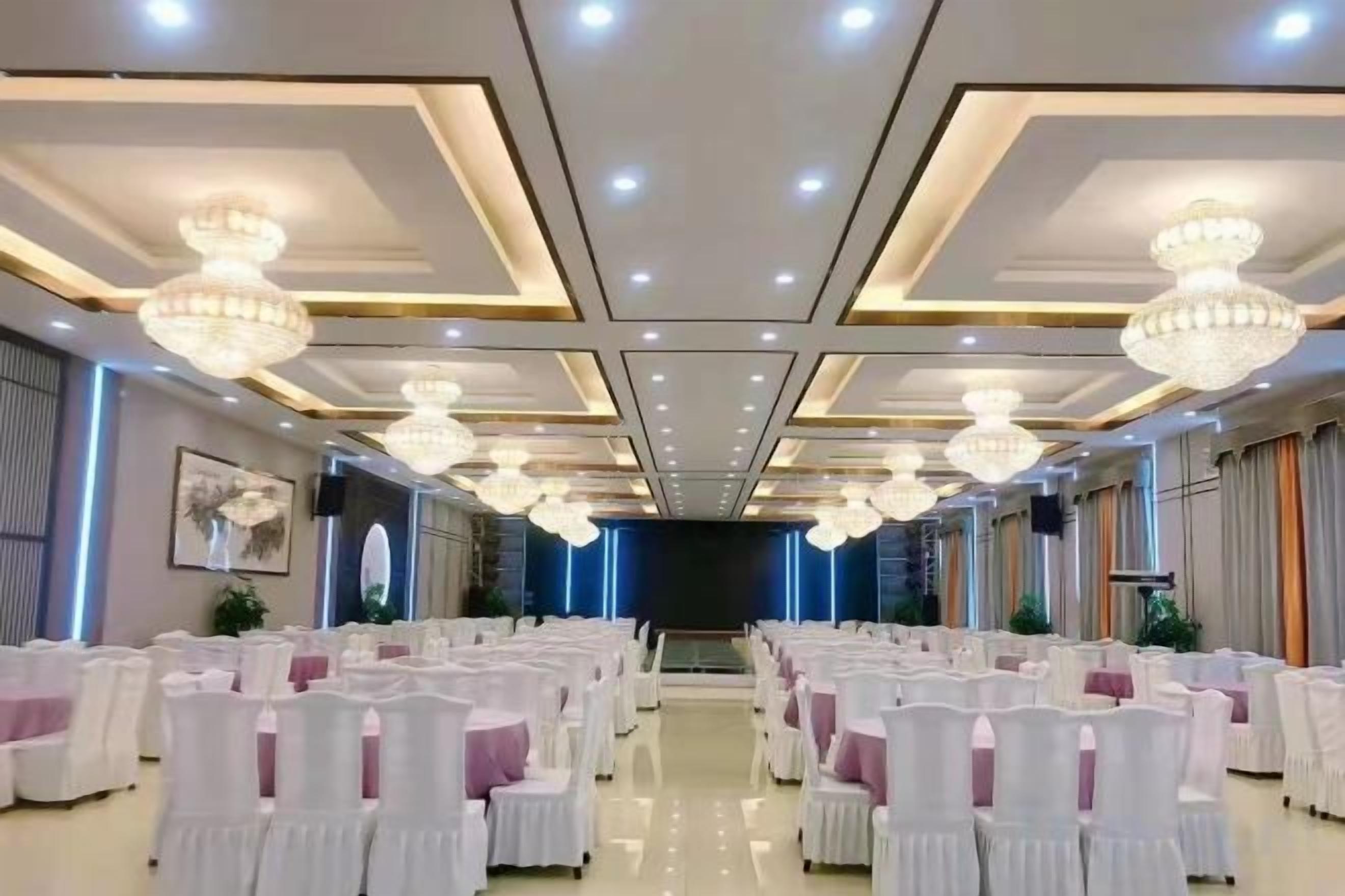 Une salle de banquet avec des rangées de chaises blanches, des lustres et une scène à l’avant.