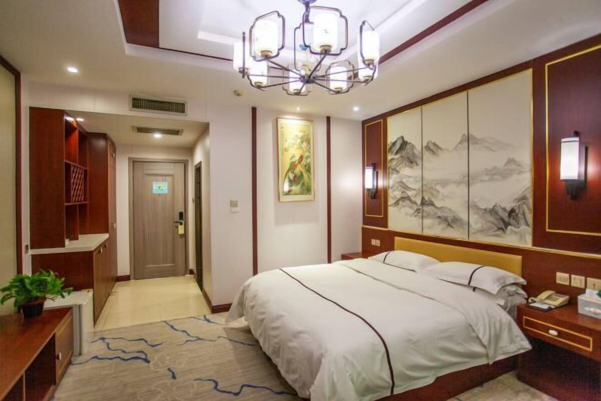 A hotel room with a large bed, a chandelier, a painting on the wall, and a desk with a plant.