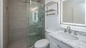 Un baño moderno con ducha de vidrio, inodoro blanco, lavamanos de mármol y un espejo grande.