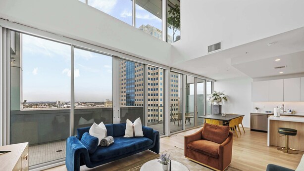 A modern living room with a blue sofa, a brown armchair, and a round coffee table. There is a large window offering a city view. - @marbella Lane - 3BR Coastal Retreat w/ Luxe Touch (Long Beach)