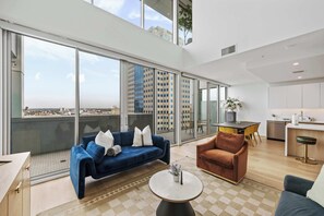 A modern living room with a blue sofa, a brown armchair, and a round coffee table. There is a large window offering a city view. - @marbella Lane - 3BR Coastal Retreat w/ Luxe Touch (Long Beach)