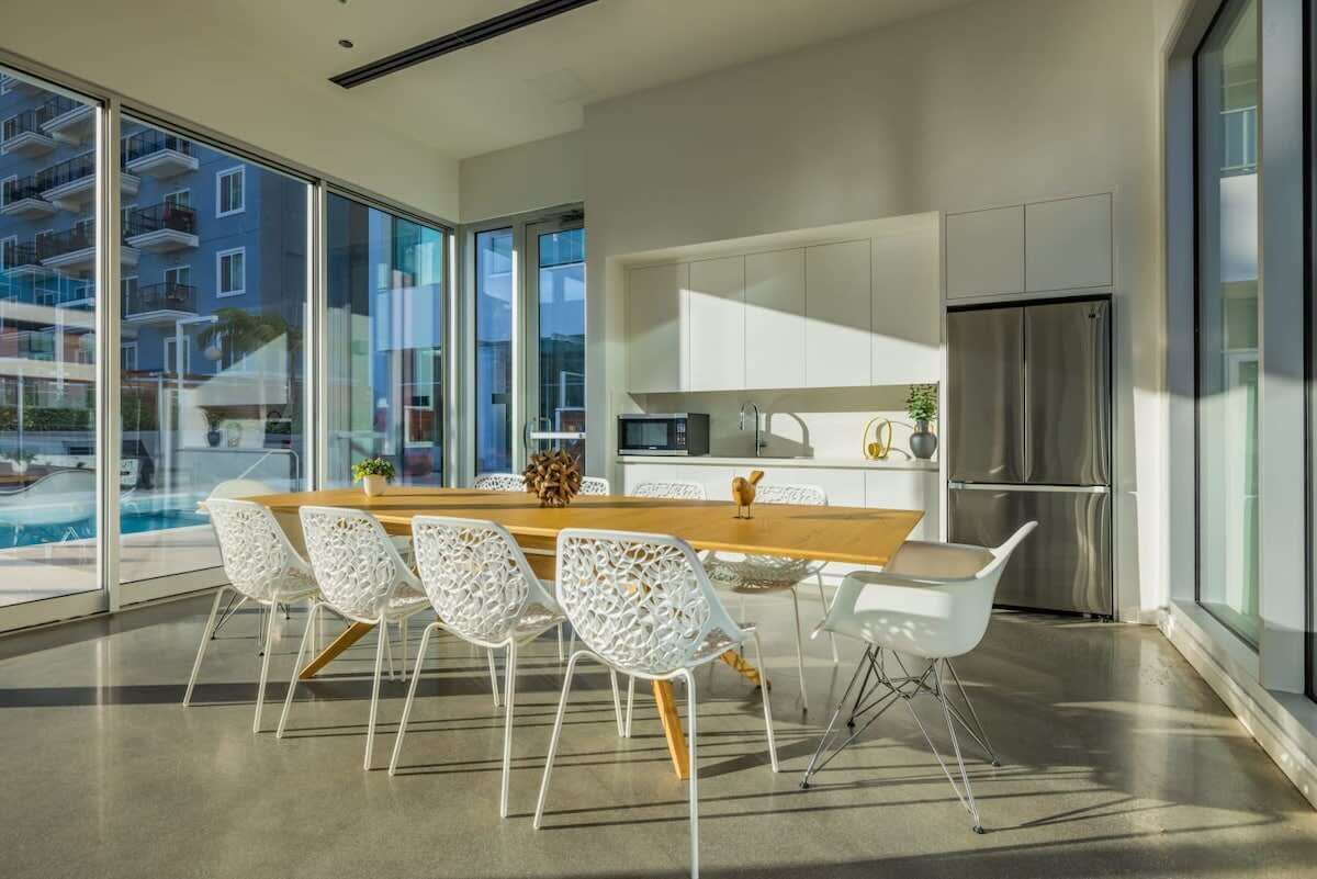 A modern kitchen with a wooden dining table and white chairs, large windows, and a view of a building outside.