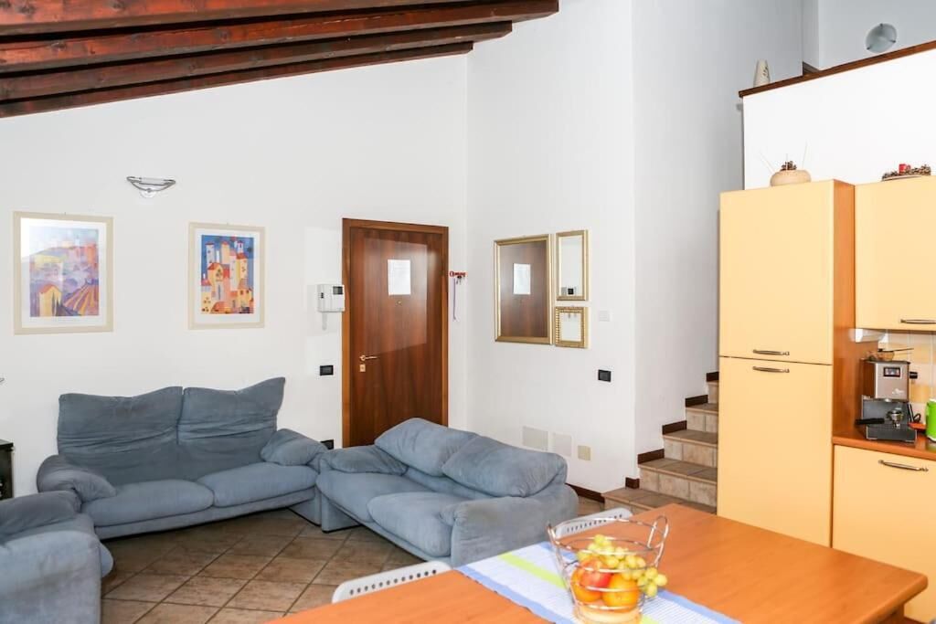 Uma sala de estar com sofá cinzento, mesa de madeira com frutas, e área de cozinha com armários e eletrodomésticos.
